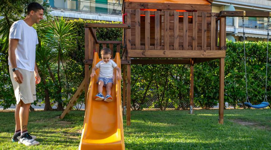 Bambino scende dallo scivolo, adulto osserva nel giardino.