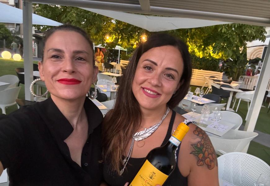 Due donne sorridenti con una bottiglia di vino in un ristorante all'aperto.