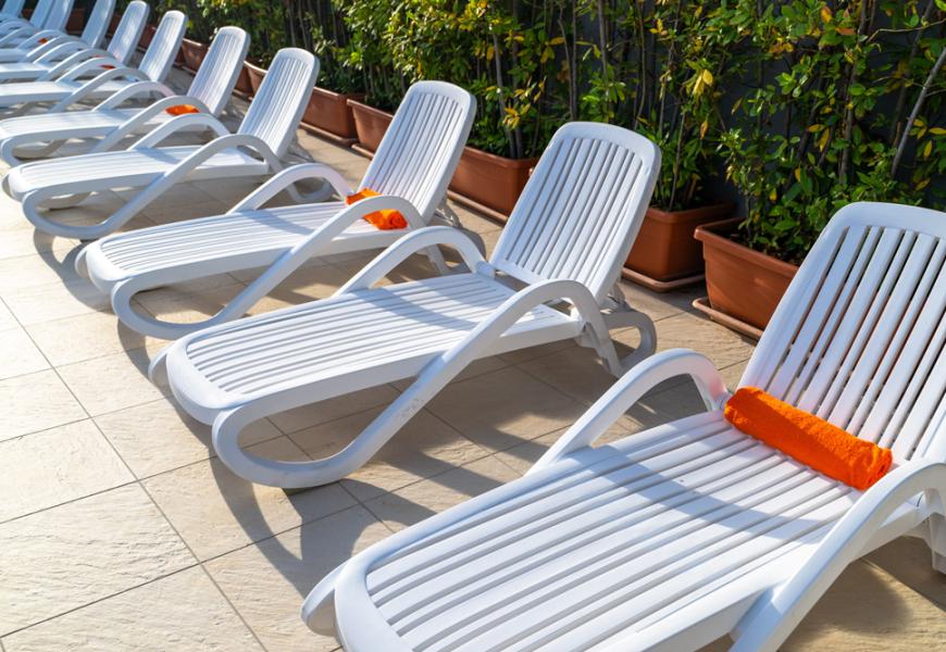 Lettini bianchi con cuscini arancioni, allineati su una terrazza soleggiata.